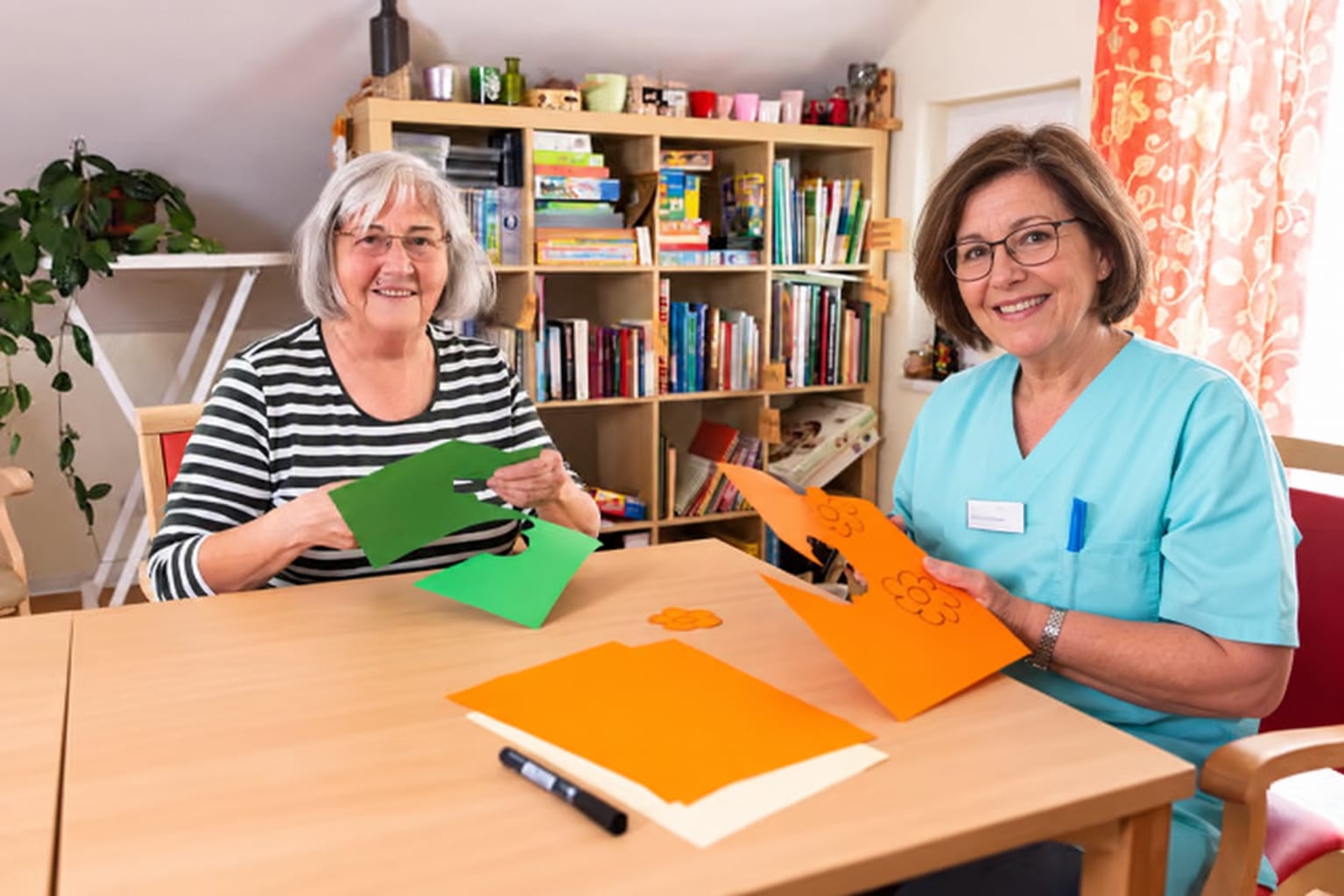 Eine Bewohnerin und eine Mitarbeiterin basteln am Tisch mit grünem und orangem Tonpapier und lächeln in die Kamera