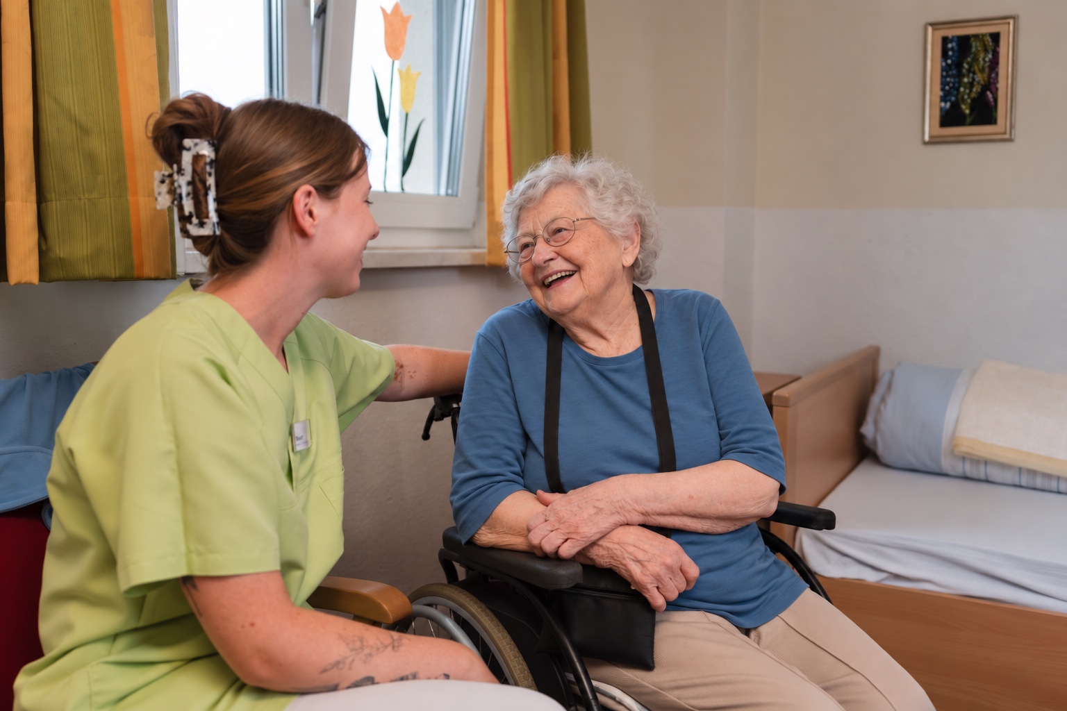 Pflegekraft und Bewohnerin im Rollstuhl lachen miteinander in ihrem Zimmer, daneben das Bett, am Fenster Tulpen aus Papier