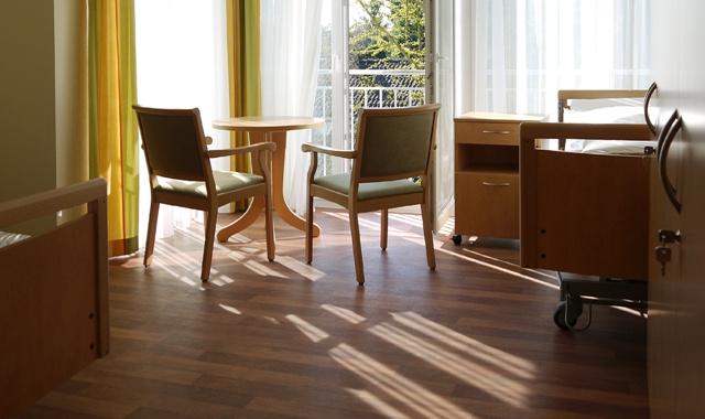 Sonniges Doppelzimmer mit je einem Pflegebett links und rechts, in der Mitte ein runder Tisch mit zwei Stühlen vor der Balkontür
