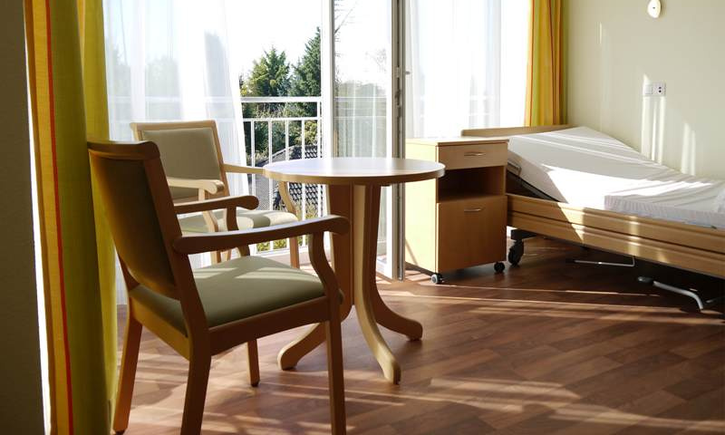 Helles Einzelzimmer mit rundem Tisch und zwei Stühlen vor der offenen Tür zum französischen Balkon, rechts Pflegebett und Nachttisch