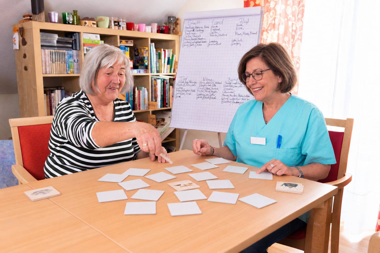 Eine Bewohnerin und eine Mitarbeiterin spielen am Tisch ein Gedächtnisspiel mit Bildkarten, dahinter ein Bücherregal und ein Flipchart mit „Stadt, Land, Fluss“