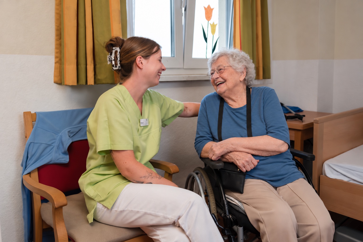 Pflegehelferin sitzt neben einer Bewohnerin im Rollstuhl, beide lachen miteinander, dahinter das Bett im Bewohnerzimmer